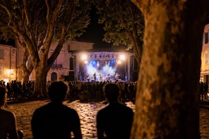 Concert en plein air la nuit sur une place de village provençal bordée de grands platanes, public en silhouette.