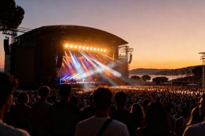 Grand stade en plein air à la tombée du jour pendant un concert d'été, foule face à une scène illuminée sur la Côte d'Azur