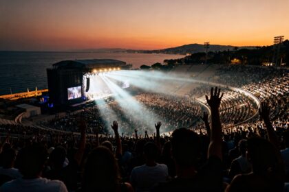 Concert en plein air dans un grand stade au coucher du soleil, public aux mains levées face à la scène et à la mer.
