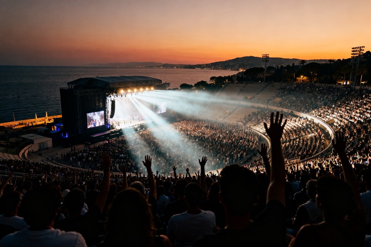 Concert en plein air dans un grand stade au coucher du soleil, public aux mains levées face à la scène et à la mer.