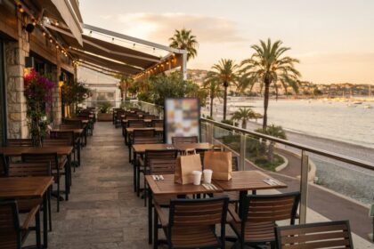 Terrasse en bois d'un restaurant face à la mer, sacs en papier kraft sur une table, palmiers et plage au coucher du soleil
