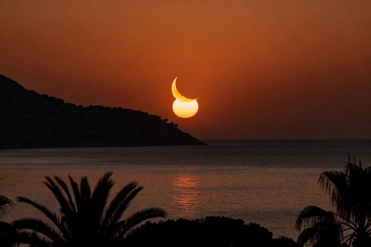 Soleil en croissant partiellement éclipsé, bas sur la mer au crépuscule, avec silhouettes de collines et de palmiers