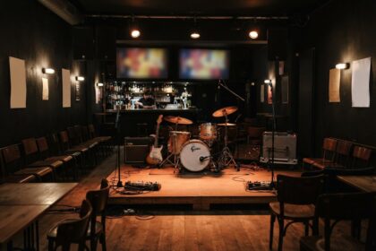 Scène en bois d'une petite salle de concert, batterie, guitare et amplis sous les projecteurs, chaises et bar en fond