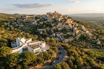 Vue aérienne d'un village provençal perché au soleil couchant, un bâtiment blanc moderne et des voitures noires en contrebas