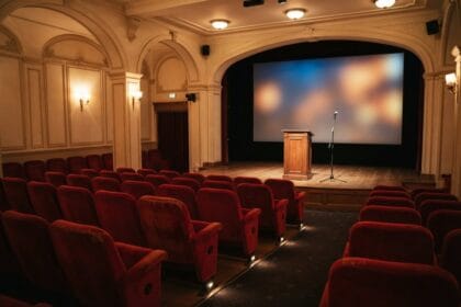 Salle de cinéma vide aux fauteuils rouges, pupitre en bois et micro devant l'écran éclairé