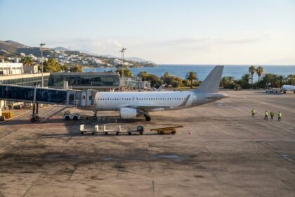 Avion de ligne blanc stationné sur le tarmac d'un aéroport méditerranéen, passerelle en place, mer et palmiers au fond