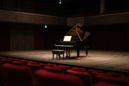 Piano à queue noir seul sur la scène d'un auditorium, tabouret et partition blanche, fauteuils rouges au premier plan