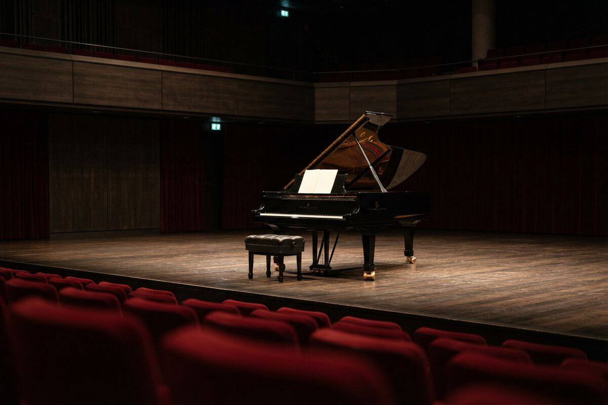 Piano à queue noir seul sur la scène d'un auditorium, tabouret et partition blanche, fauteuils rouges au premier plan