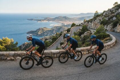 Trois cyclistes en tenue bleue montent une route en corniche au-dessus d'une baie méditerranéenne