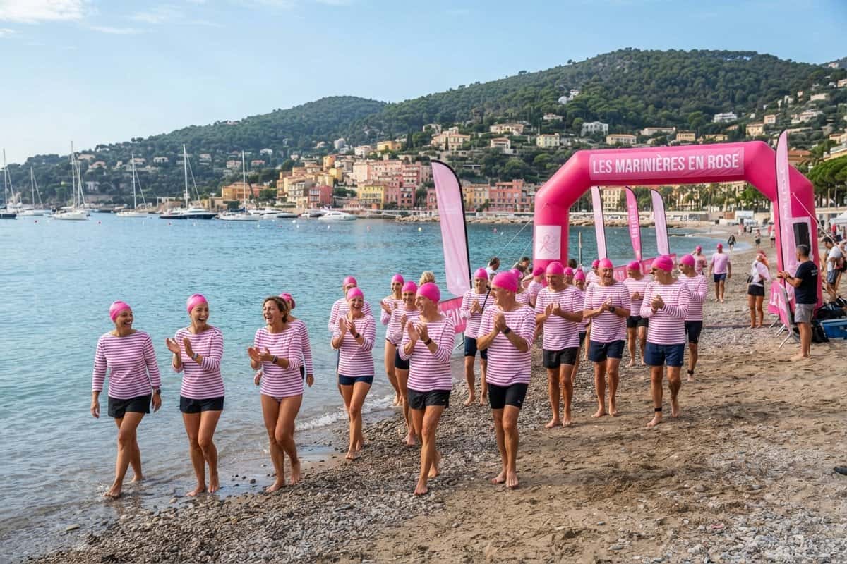 Nageurs en marinière rose et bonnet rose sur une plage de galets, arche gonflable rose, rade et collines en fond
