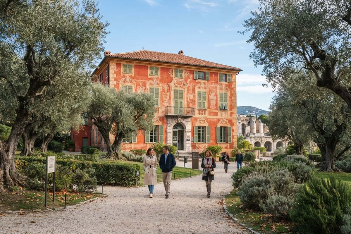 Villa ocre rouge aux volets verts entourée d'oliviers, des visiteurs marchant sur une allée de gravier